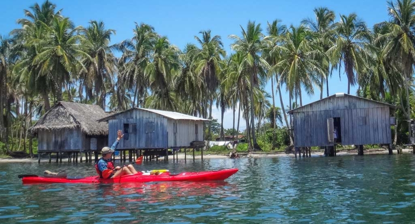 A person in a red kayak paddles past three stilt houses and a row of palm trees. 